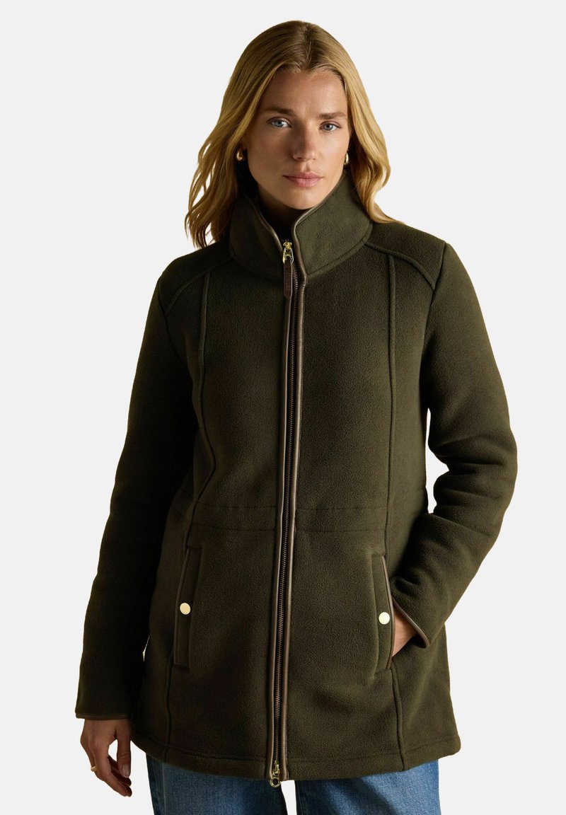 Kobieta w ciemnozielonej polarowej kurtce z kołnierzem stojącym, zamkiem z przodu i kieszeniami na ręce, pozująca na jednolitym jasnym tle.