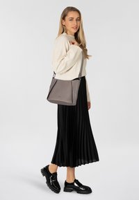 Bolso de mano gris texturizado con una correa plateada, llevado cruzado. Modelo en un suéter crema y falda negra con plisados, de pie sobre un fondo blanco.