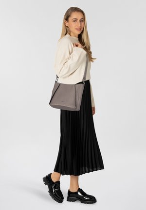 Graue, strukturierte Handtasche mit einem silbernen Tragegurt, im Crossbody-Stil getragen. Model in einem cremefarbenen Pullover und schwarzem, plissiertem Rock, auf einem weißen Hintergrund stehend.