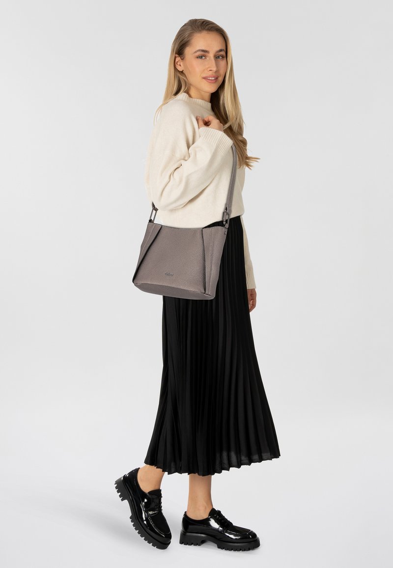 Bolso de mano gris texturizado con una correa plateada, llevado cruzado. Modelo en un suéter crema y falda negra con plisados, de pie sobre un fondo blanco.
