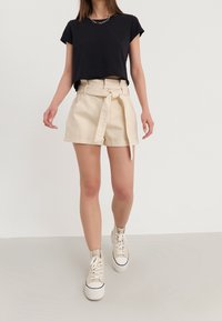 Černé zkrácené tričko, béžové šortky s vysokým pasem a předním zavazováním a kapsami, doplněné béžovými vysokými teniskami na světlem pozadí.