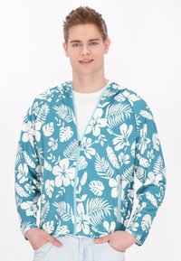 Huvtröja i teal med vita blommor och bladmönster. Utvecklad med dragkedja, långa ärmar och ett lätt, strukturerat tyg.