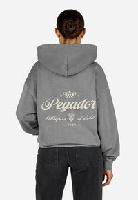 Sivá prešívaná mikina s veľkým strihom a sťahovacou kapucňou, na zadnej strane so žltým potlačeným textom "Pegador" a dekoratívnymi prvkami. Bavlnený materiál.
