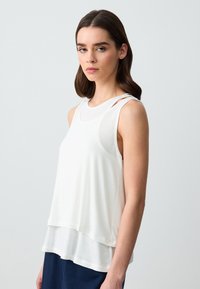 Top bianco a strati senza maniche con scollo rotondo e sottili spalline. Tessuto morbido e leggero con una vestibilità ampia.