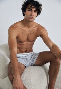 Plikas jaunas vyras su garbanotais plaukais sėdi ant tekstūruoto balto kėdės, vilkėdamas šviesiai pilkus dryžuotus Calvin Klein šortus.