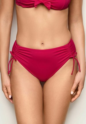 Žena nosící červené bikiny s vysokým pasem a bočními zavazovadly a odpovídající horní díl bikin, který odhaluje střed těla a ruce odpočívají podél stran.