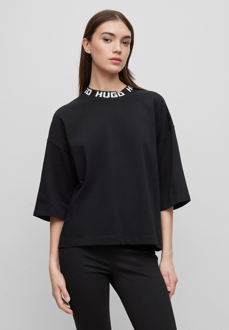 T-shirt nera oversize con collo alto e il testo bianco "HUGO". Materiale in cotone morbido con spalle scese e vestibilità rilassata.