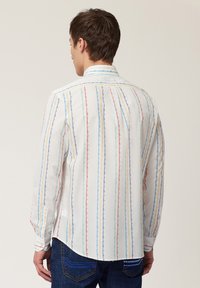 Witte blouse met lange mouwen en verticale meerkleurige strepen (rood, blauw, geel) en een knoopsluiting bij de kraag. De stof lijkt glad en lichtgewicht te zijn.