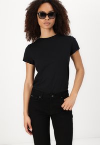 Sieviete ar cirtainiem matiem, valkājot melnas saulesbrilles, melnshort-sleeve t-kreklu un melnas bikses, stāv uz baltas fona.