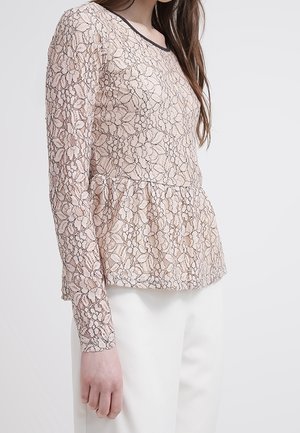 Femme portant un haut en dentelle beige à manches longues avec des motifs floraux et une taille peplum, associé à un pantalon blanc, sur un fond uni.