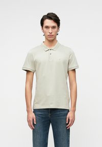 Világosszürke pólóing gallérral, két gombbal, rövid ujjakkal és diszkrét logóval. Sötétkék farmernadrággal viselve, minimalista design.