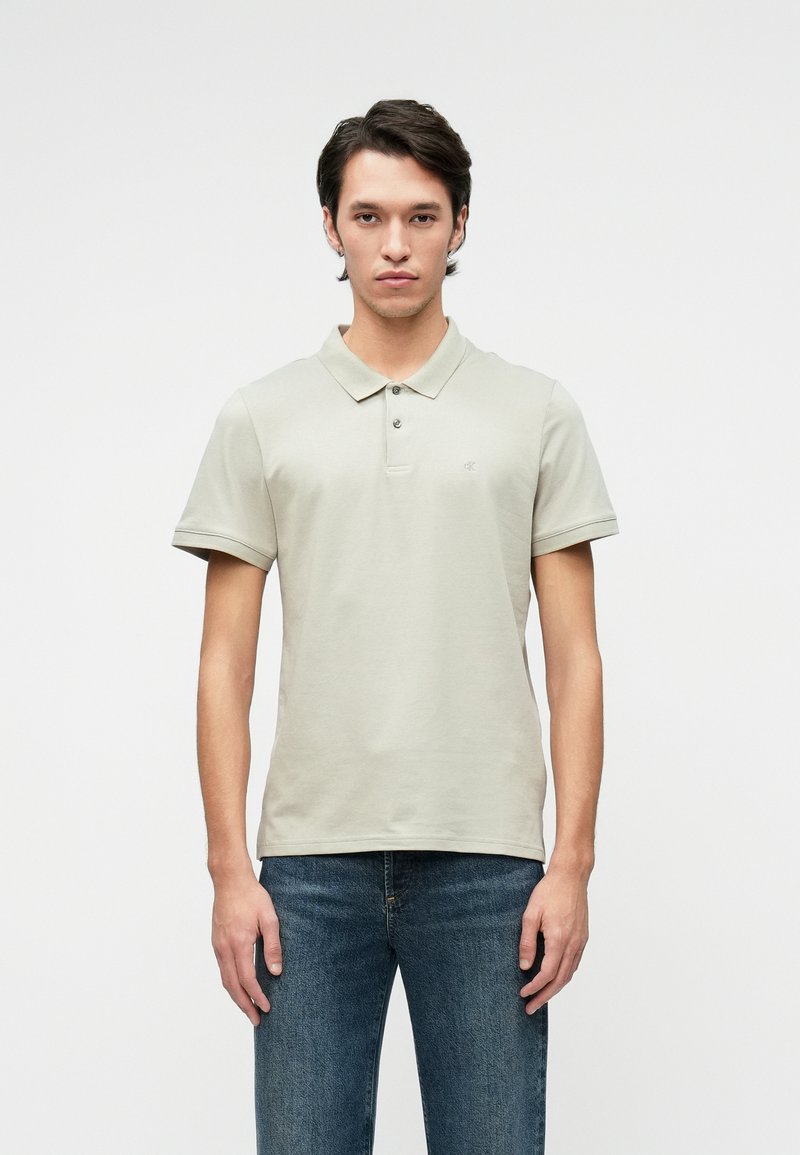 Šviesiai pilkas polo marškinėlis su apykakle, dviem mygtukais, trumpomis rankovėmis ir subtiliu logotipu. Dėvima su tamsiai mėlynomis džinsomis, minimalistiško dizaino.