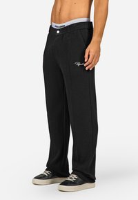 Fekete, gofrés mintájú nadrág, testre szabott derékkal és elasztikus derékszalaggal, oldalzsebekkel és egy kis fehér logóval a bal lábszáron.