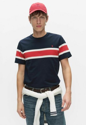 Młody mężczyzna w granatowym t-shircie z czerwonymi i białymi paskami, czerwonej czapce, niebieskich dżinsach i białym swetrze związanym wokół talii.