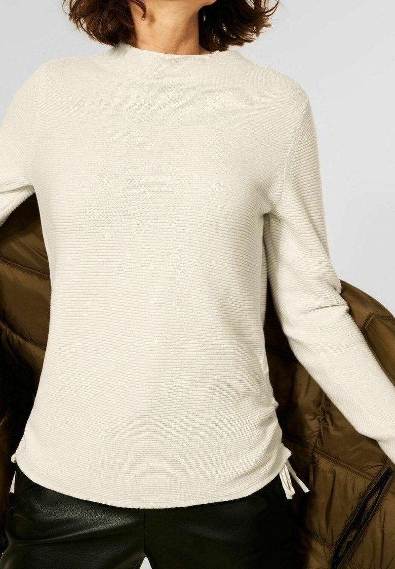 Maglione a coste di colore crema con maniche lunghe, dotato di un collo alto e lacci laterali, abbinato a un giubbotto imbottito marrone.