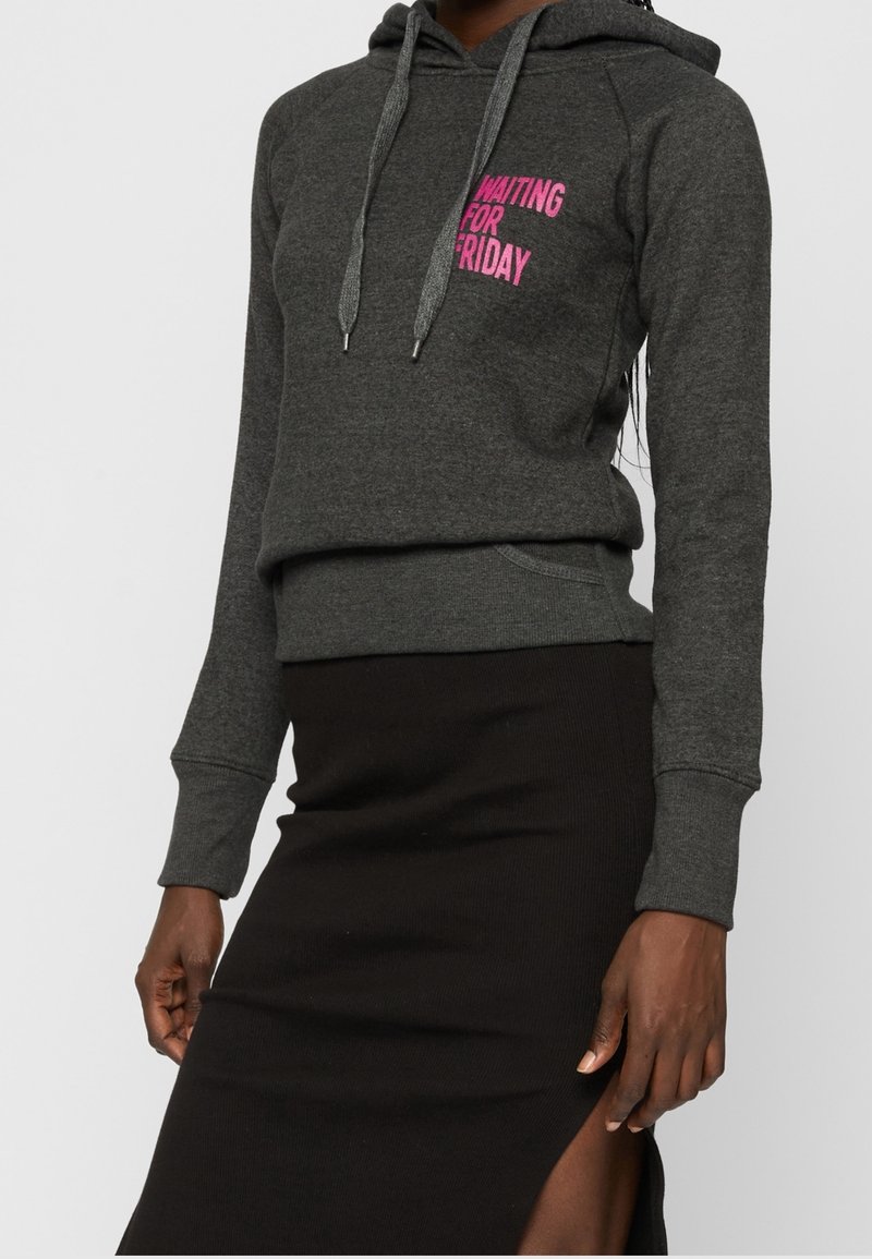 Personne portant un sweat à capuche gris foncé avec un texte rose "WAITING FOR FRIDAY" et une jupe noire avec une fente latérale.
