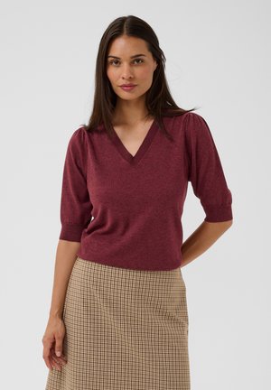 Femme aux longs cheveux foncés portant un haut bordeaux en V avec des manches aux coudes et une jupe beige à carreaux, debout devant un fond uni.