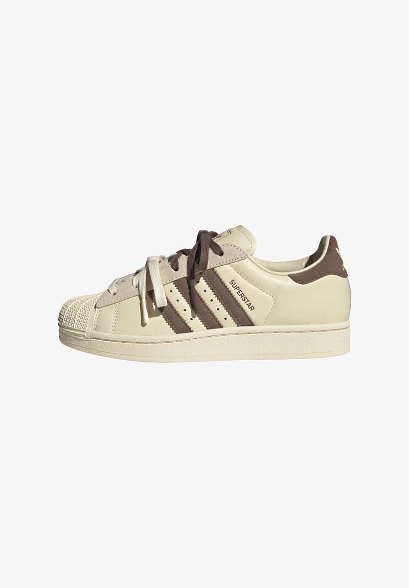Κλασικό sneaker χαμηλού πάνω μέρους σε κρεμ χρώμα με καφέ ρίγες από σουέντ και κορδόνια, καουτσούκ μύτη σε σχήμα κελύφους, και κείμενο "SUPERSTAR" στο πλάι.