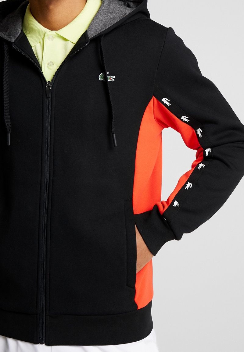 Svart och orange hoodie med dragkedja fram, grå foder i huvan, kängurufickor och vita logodetaljer på ärmarna. Mjuk, sportig design.