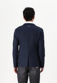 Navy blazer with a smooth texture, single vent at the back, and logo detail near the collar, paired with dark gray jeans.