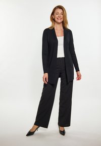 Cardigan nero aperto realizzato in tessuto morbido, abbinato a una maglietta bianca e pantaloni neri a gamba larga. Gli accessori includono orecchini a cerchio.