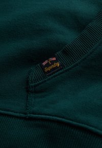 Sudadera verde oscura con puños y dobladillo acanalados, con una pequeña etiqueta del logo de Superdry cosida en el lateral. Textura suave, diseño casual.