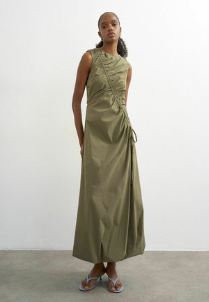 Kvinde iført lang, ærmeløs kjole i olivengrøn med rynket diagonalt mønster og lyseblå hælede sandaler, stående mod en ensfarvet væg.