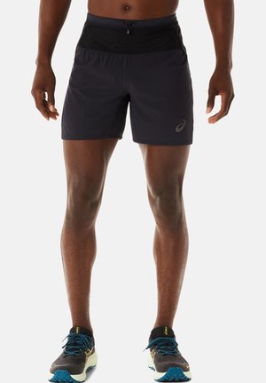 Mann trägt schwarze Sportshorts mit Reißverschlusstasche und schwarze Laufschuhe mit blauen Schnürsenkeln, steht vor einem einfarbigen Hintergrund.