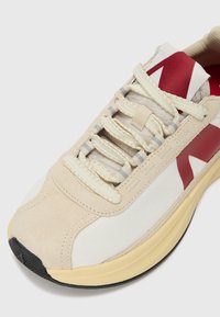 Beige and white sneaker with a smooth suede and textured fabric upper, red accents, and a thick yellow rubber sole. Features laces and a padded collar.