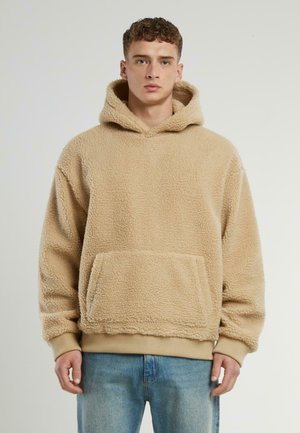 Beige fleece hoodie with a textured surface, large hood, and front pocket. Features ribbed cuffs and hem. Paired with light blue jeans.