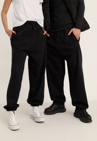 Black sweatpants with a relaxed fit, elastic cuffs, and drawstring waist. Worn with contrasting white and black sneakers.