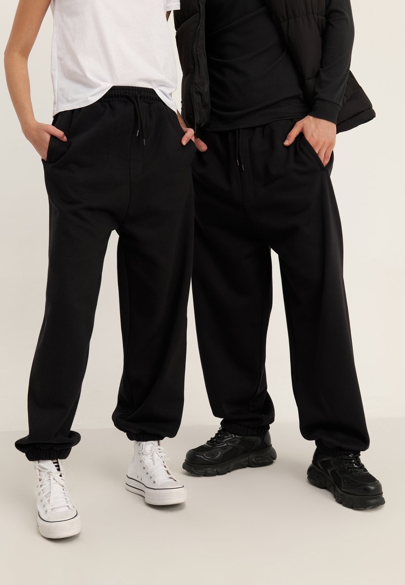 Black sweatpants with a relaxed fit, elastic cuffs, and drawstring waist. Worn with contrasting white and black sneakers.