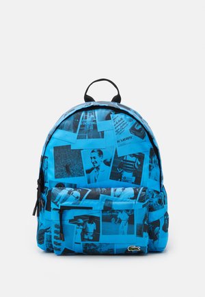 Blå rygsæk med sort-hvide vintage-fotos og Lacoste-logo med en grøn krokodille på den nederste forlomme.