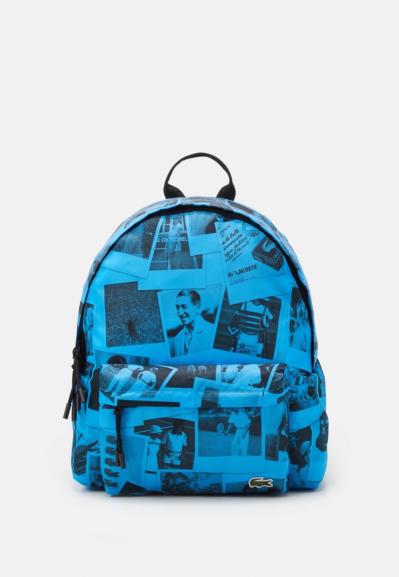 Sac à dos bleu avec des photos vintage en noir et blanc et le logo Lacoste avec un crocodile vert sur la poche avant inférieure.