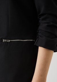 Černá bunda s elegantním designem. Má kapsu na zip a na rukávech nařasené detaily. Měkká textura látky, přiléhavý střih.