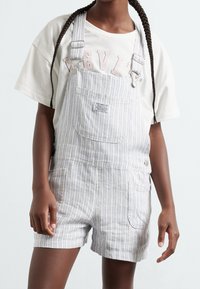 Gestreifte Latzhose aus einer Baumwollmischung, hellblau und weiß, mit Tasche vorne und Knopfverschlüssen. Getragen über einem beigen T-Shirt mit Grafikdruck.