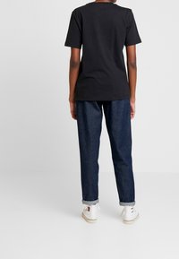 Camiseta de algodón negra de manga corta, combinada con pantalones de mezclilla oscuros enrollados y zapatillas blancas, vista desde la parte de atrás.