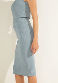 Žena ve světle modrém žebrovaném topu bez rukávů a odpovídající úzké žebrované sukni stojí na neutrálním pozadí.