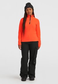 Sudadera de forro polar de color naranja brillante con cremallera de cuarto, combinada con pantalones cargo negros que tienen bolsillos y un corte holgado. Gorro negro en la cabeza.