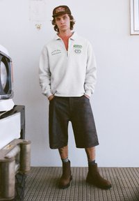 Felpa grigia in pile con cerniera fino a metà, logo verde, pantaloncini scuri e stivaletti marroni alla caviglia. Indossata con un cappellino nero con logo.