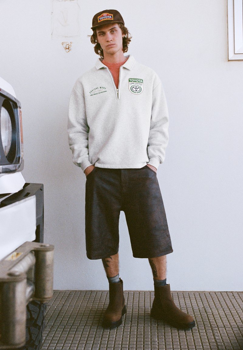 Felpa grigia in pile con cerniera fino a metà, logo verde, pantaloncini scuri e stivaletti marroni alla caviglia. Indossata con un cappellino nero con logo.