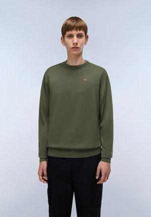 Giovane uomo che indossa un maglione a maniche lunghe di colore verde oliva con una piccola emblema a forma di bandiera sul petto e pantaloni cargo neri, in piedi contro uno sfondo chiaro e semplice.