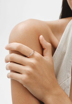 Anillo de plata con tres pequeñas gemas transparentes, usado en una mano que reposa sobre un hombro desnudo; piel suave y fondo neutro.