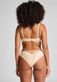 Frau mit lockigem Haar, die beiges Lingerie mit Kreuzdetail auf dem Rücken trägt, steht vor einem einfarbigen hellgrauen Hintergrund.