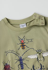 Khakinvärinen puuvillapaita, jossa on printattuja värikkäitä kovakuoriaisia, pyöreä pääntie ja nepparit yhdessä olkapäässä helppoa pukemista varten.