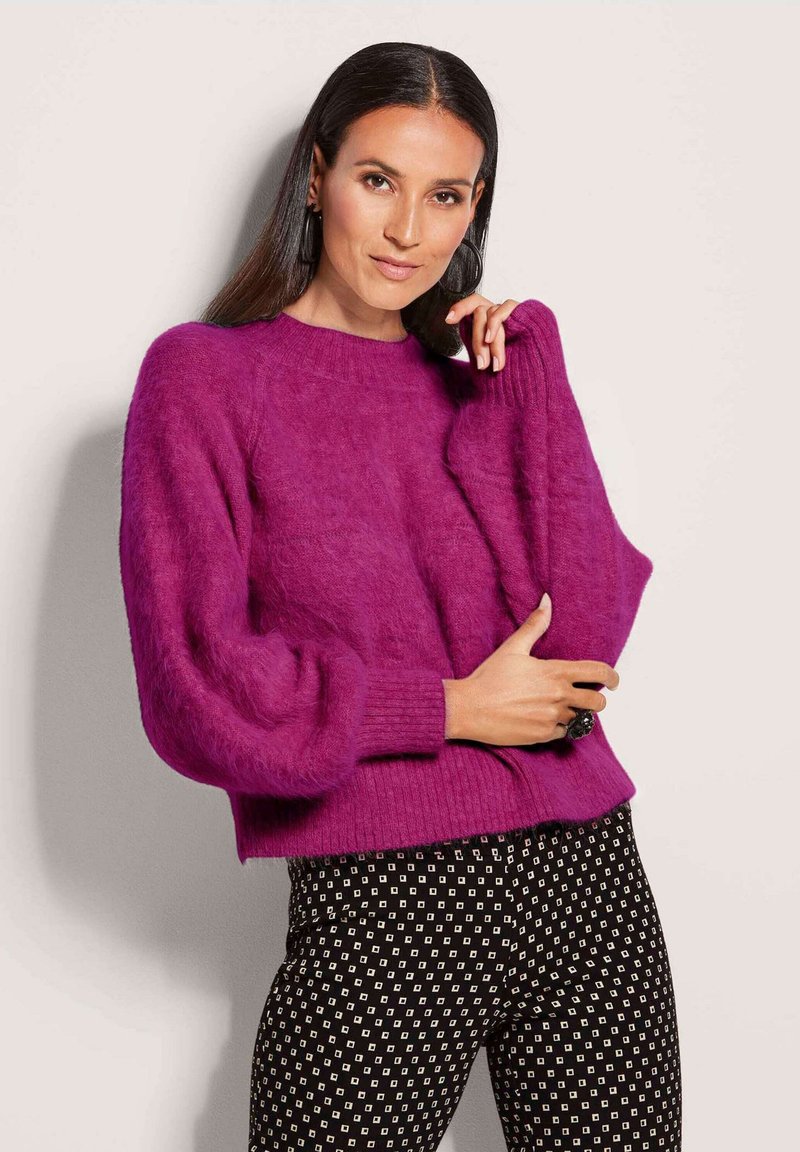Flauschiger lila Pullover mit gerippten Bündchen und Saum, lockere Passform und runder Ausschnitt, kombiniert mit schwarzen Hosen mit einem weißen geometrischen Muster.