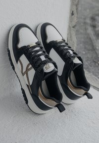 Sneakers in bianco e nero con upper in pelle, pannelli texture, dettagli dorati e lacci a trama, caratterizzati da un design a profilo basso e colletto imbottito.