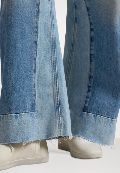 Τζιν καμπάνα σε μπλε χρώμα με διχρωμικό σχέδιο, διακοσμημένο με ξεφτισμένο τελείωμα και απαλή υφή. Συνδυασμένο με ανοιχτόχρωμα παπούτσια.