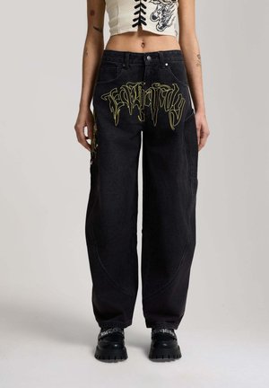 Person wearing black wide-leg jeans with yellow gothic text on front, paired with black chunky shoes and a white lace-up crop top.