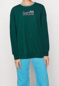 Felpa verde con collo a coste, con il logo "Levi's" ricamato in nero. Abbinata a pantaloni in velluto a coste azzurri.